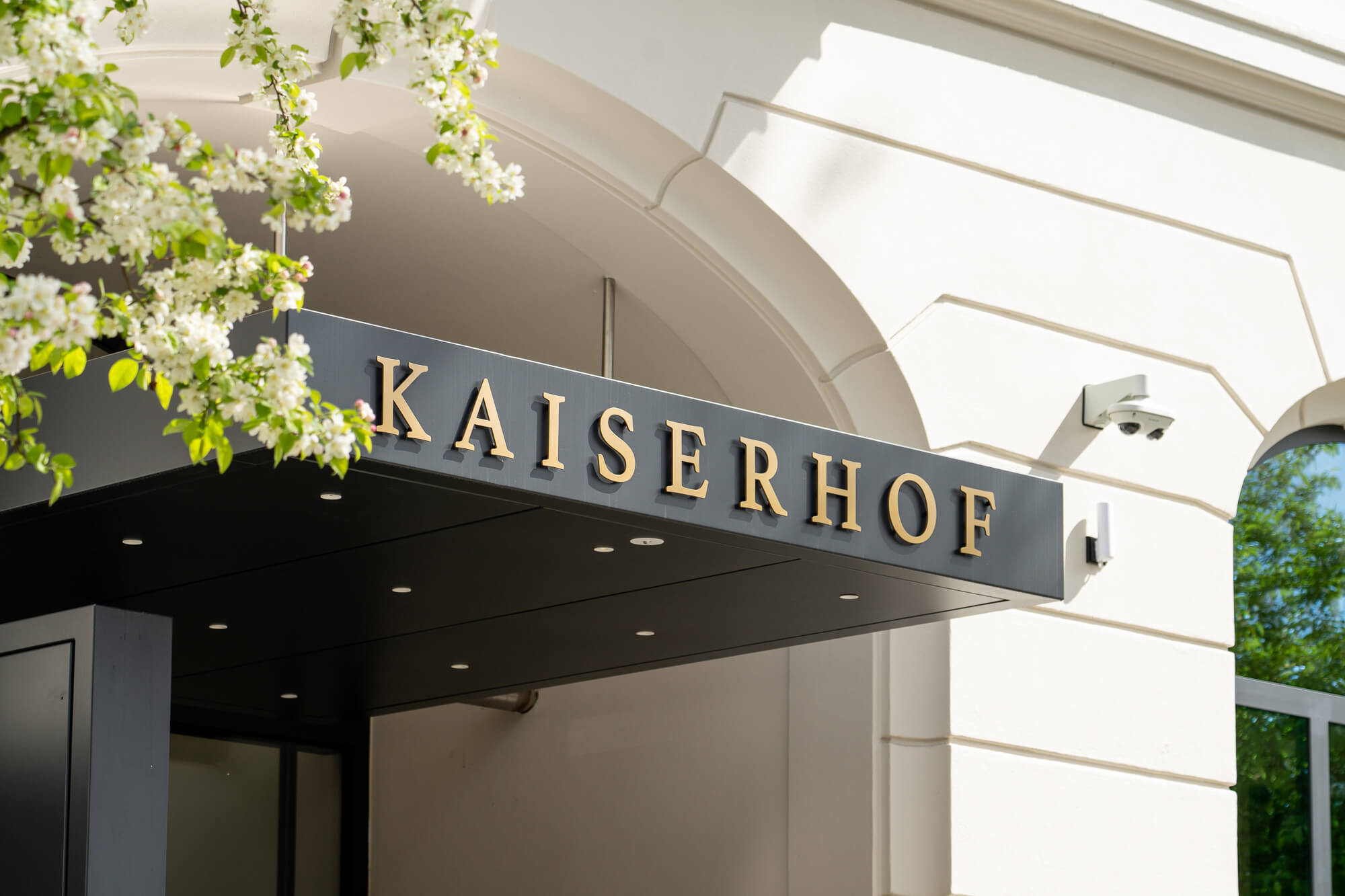 Dachelement mit Kaiserhof-Schriftzug und blühendem Baum am Entrée des Hotels