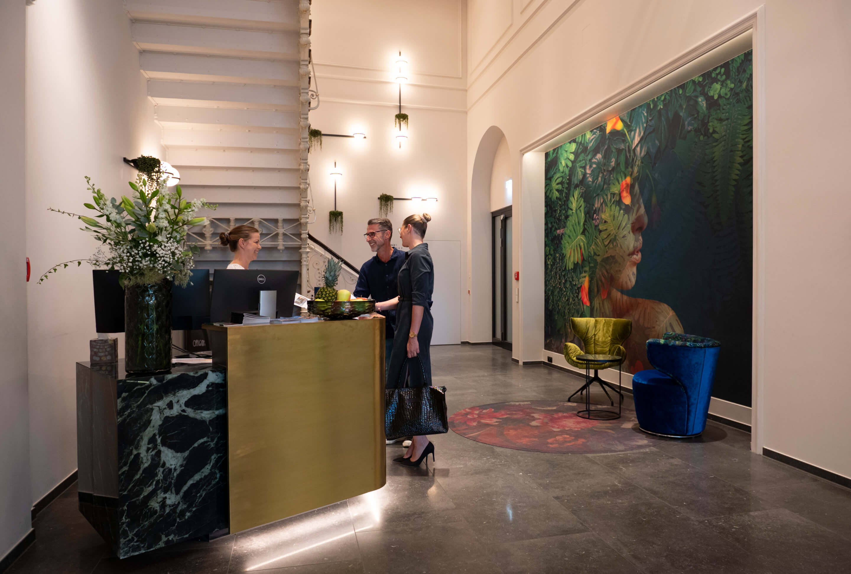 Rezeption eines stilvollen Boutiquehotels mit Gästen beim Check-in, moderner Einrichtung, großer Wandkunst und Treppenaufgang im Hintergrund